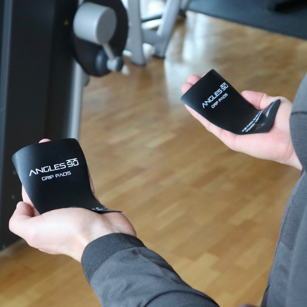 A pair of hands holding black A90 Grip Pads in a gym setting, preparing for a workout with the POWER+ GRIP Method.
