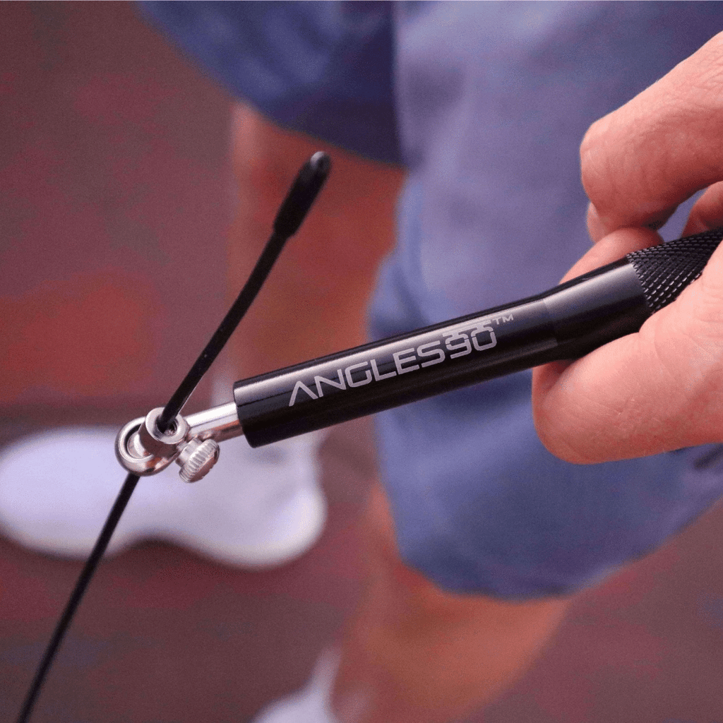 A close-up of a person's hand holding an A90 Jump Rope with in-ear headphones attached, against an outdoor training blurred background.