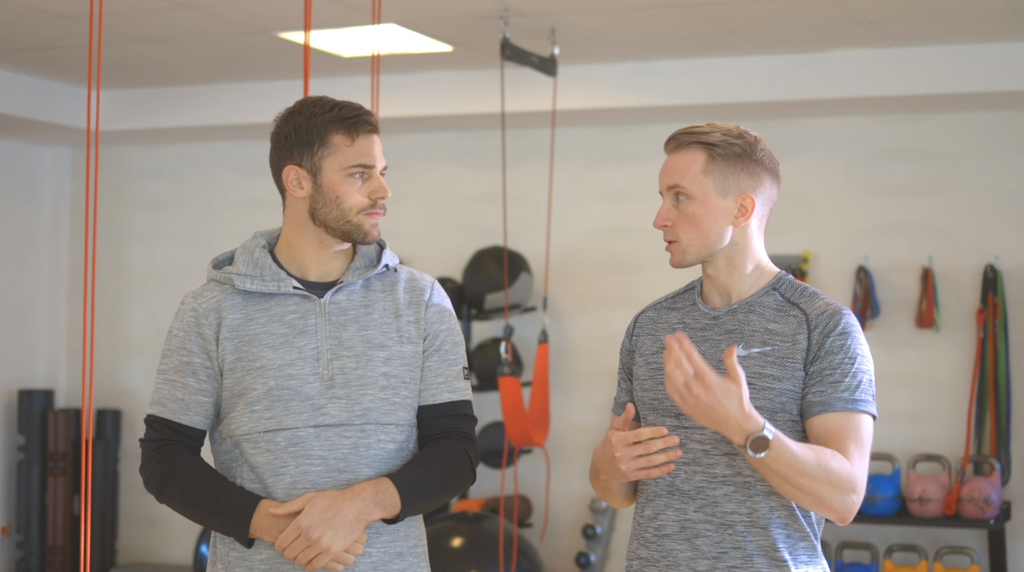 Two men having a conversation in front of an A90 Prehab: Self-Screening & Corrective Exercises (Video Course) in a gym equipped with various fitness gear.