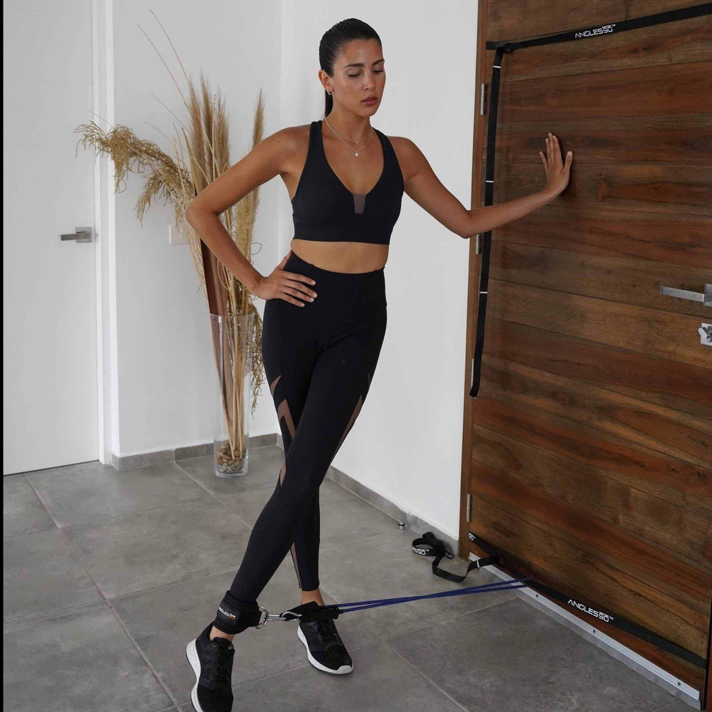 A woman in sportswear exercises with A90 Ankle Straps by a wooden door, focused on her fitness routine.