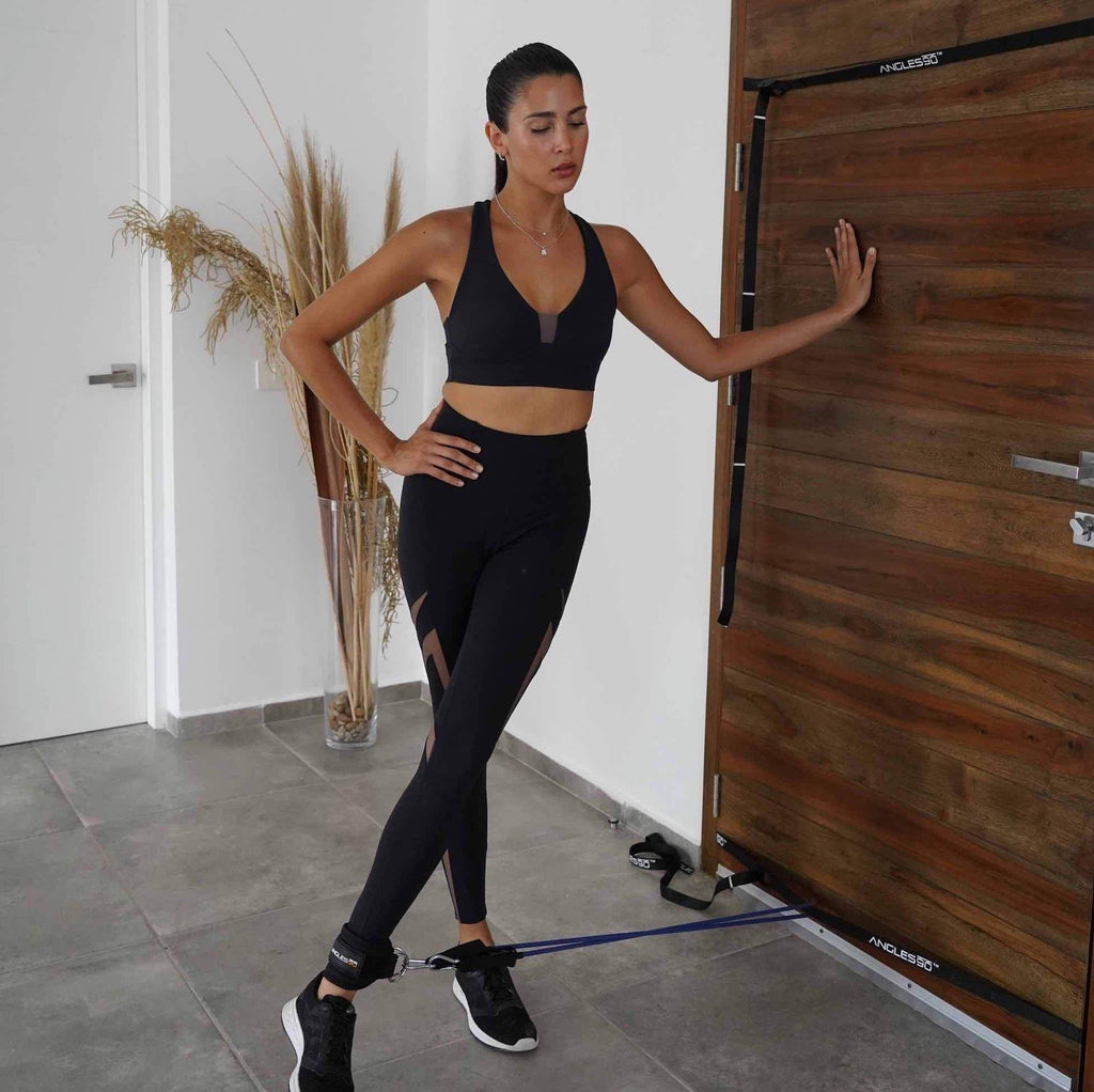A focused woman in athletic wear performing a workout with the A90 Leg Day Set near a wooden door.